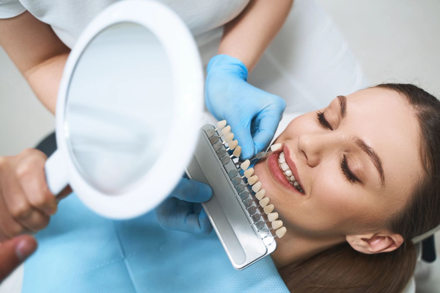 Dental professional comparing tooth shade with a guide and magnifying mirror on a smiling patient.