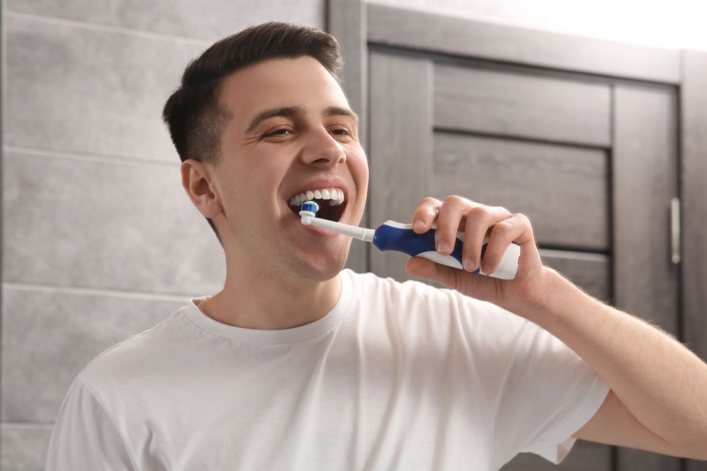 A smiling young man brushes his teeth with an electric toothbrush.