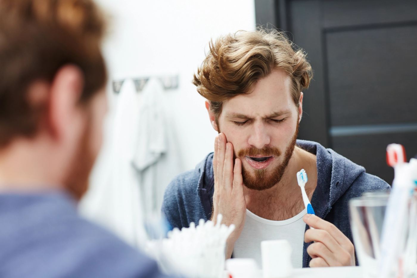 Man with toothache holding jaw and toothbrush in bathroom mirror.