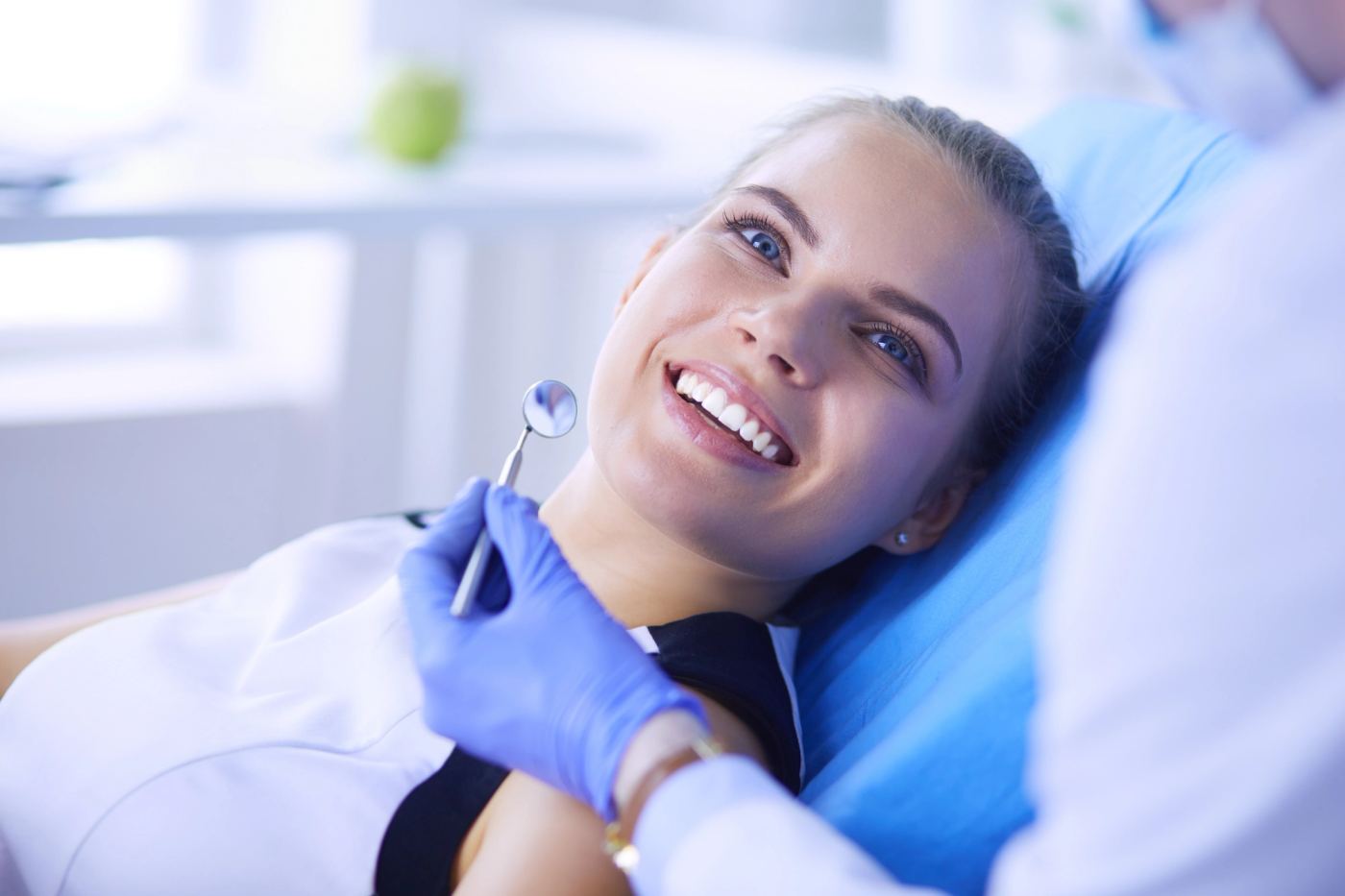A smiling woman in a dentist's chair with a gloved hand holding a dental mirror.