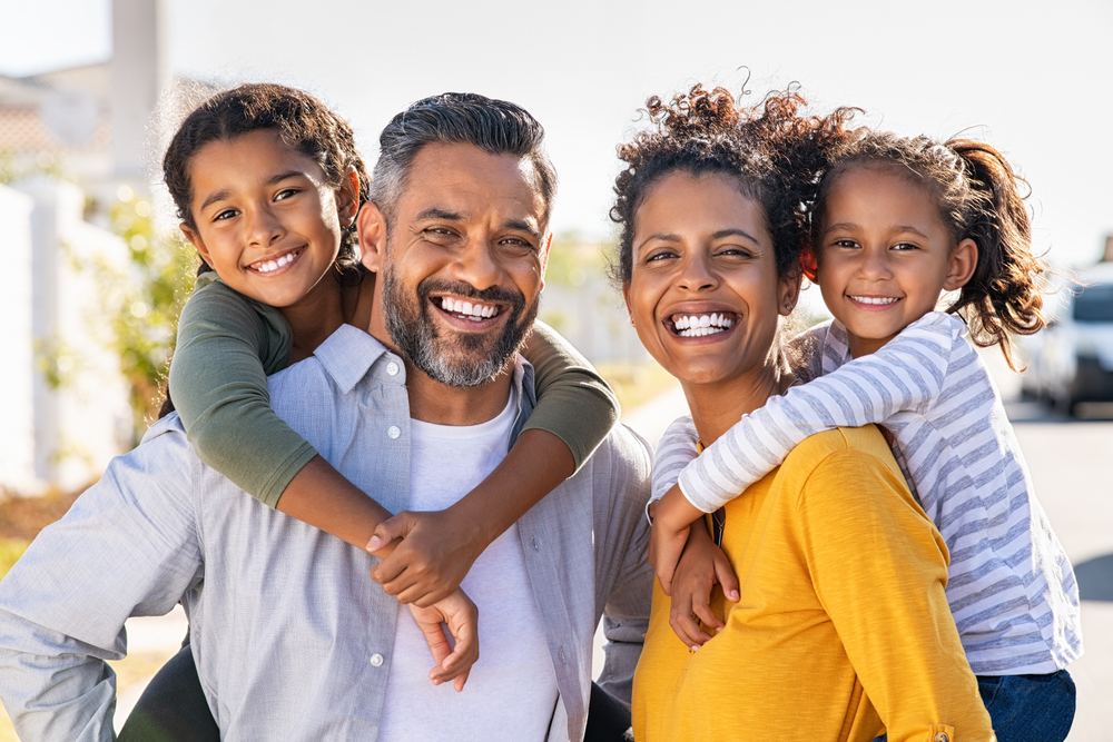 A smiling family of four, with two adults giving two children piggyback rides.