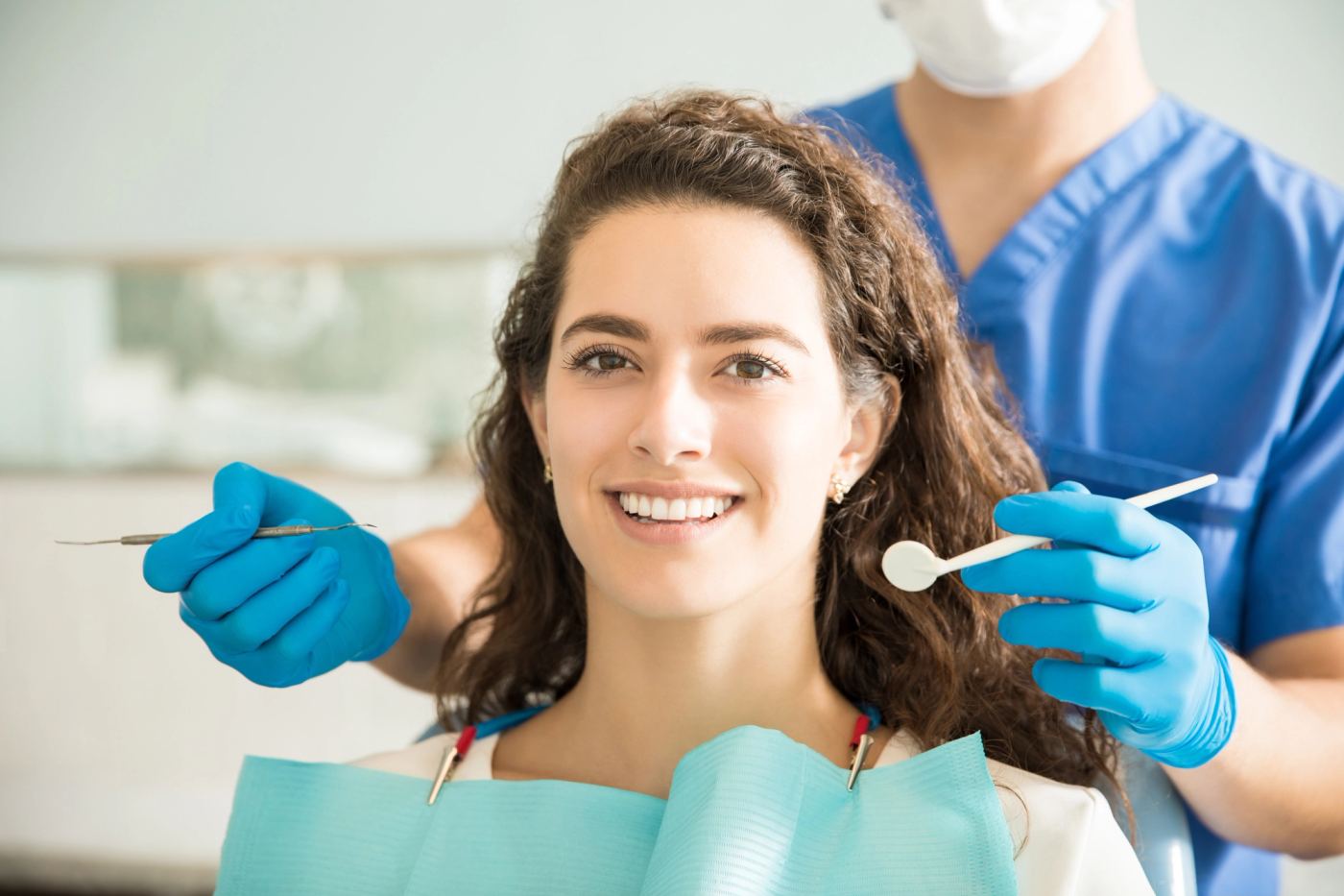 A smiling woman sits in a dental chair with a dental professional holding tools behind her.