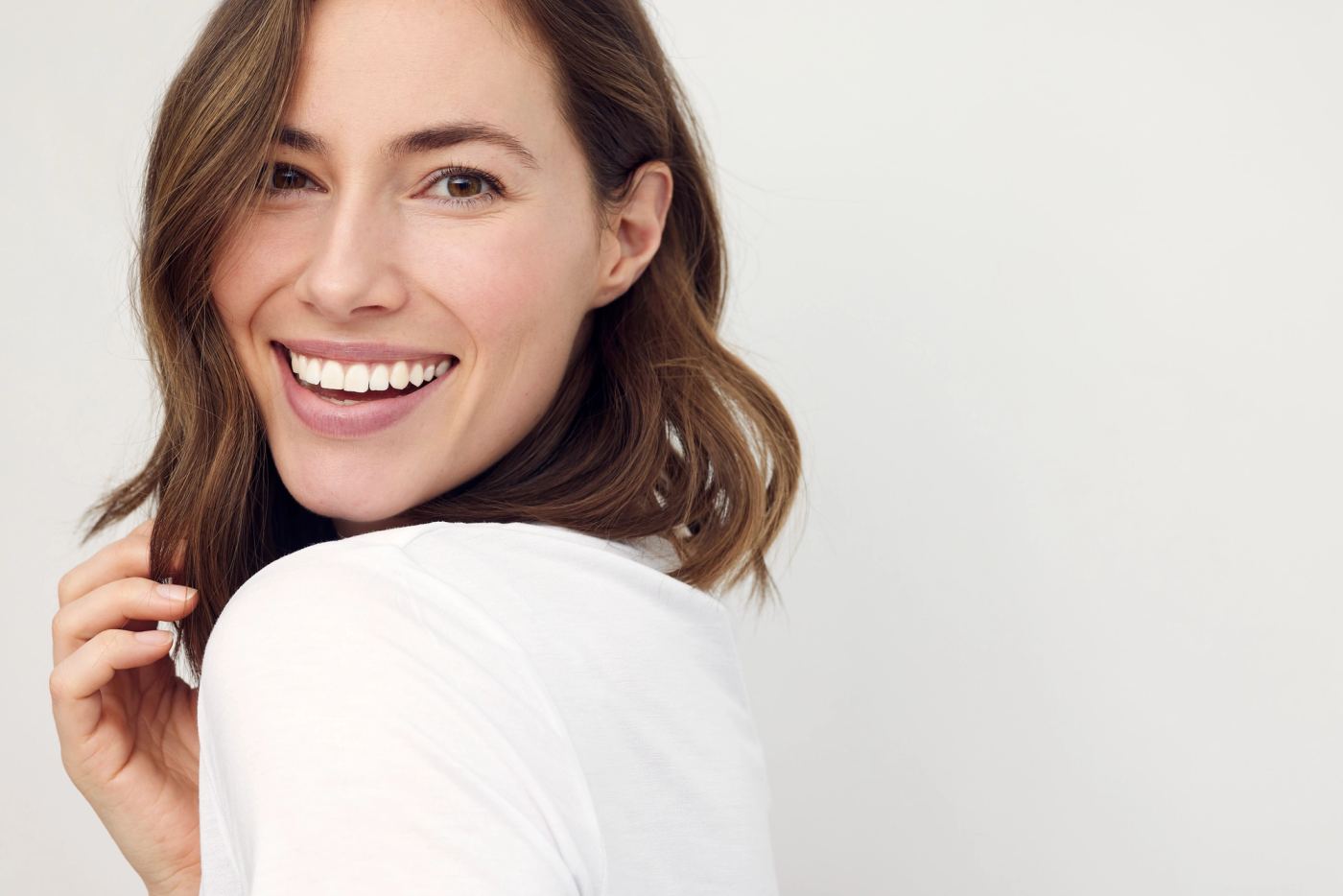 A smiling woman with wavy brown hair looks over her shoulder, wearing a white shirt.