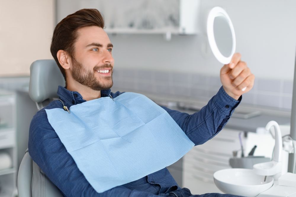 Smiling man in a dental chair, wearing a bib, looks at his reflection in a mirror.