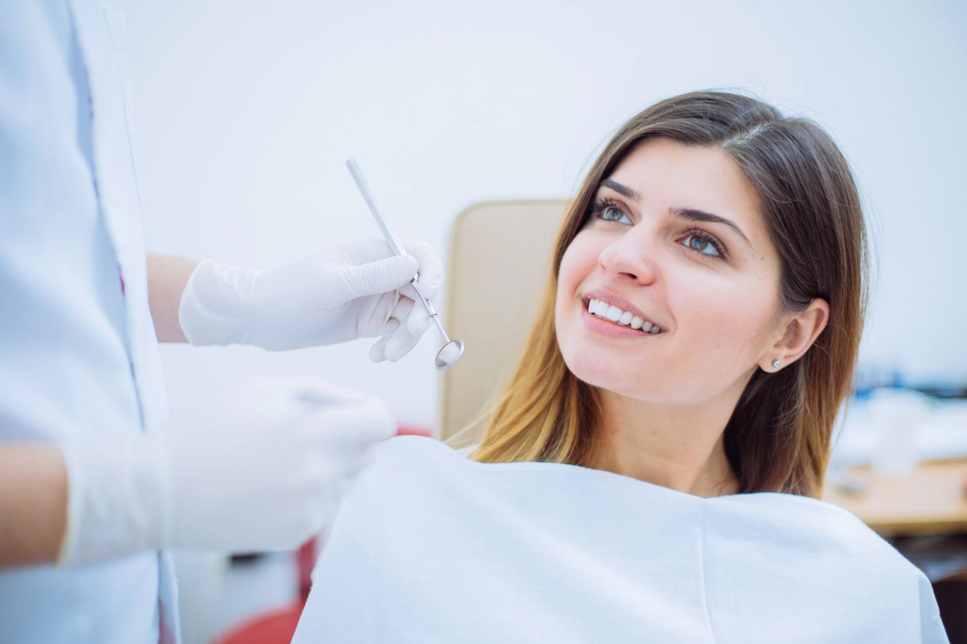 A smiling woman in a dental chair looks up at a dentist holding a dental mirror.